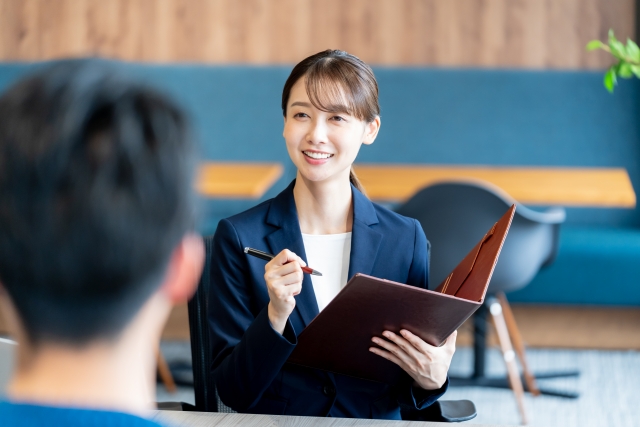 商談スペースで、スーツを着た女性がファイルを片手に説明している。手前には男性の後ろ姿が写っている。