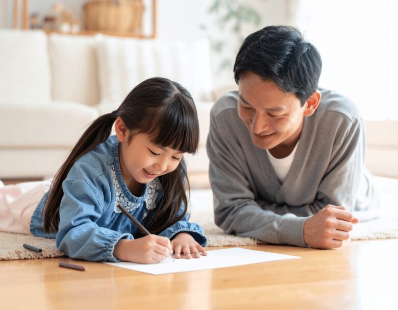 リビングに寝そべってお絵描きをしている女の子と、それを見守るお父さん