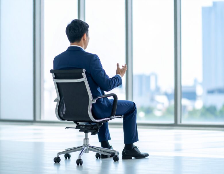 日本人男性の営業マンの写真風素材。スーツ着用の清潔感、信頼感のある風貌。明るいオフィス内の椅子に腰かけ、手振りを交えて話している様子で、腰から上のアップの斜め後ろ姿
