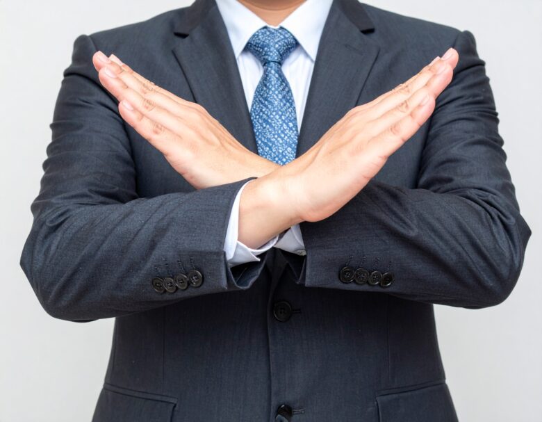 スーツを着た男性が、両手を交差させて「バツ」の形を作る(拒否・否定のポーズ)。手首や前腕で×印を作っている構図。顔は写っておらず、手元のアップ。
