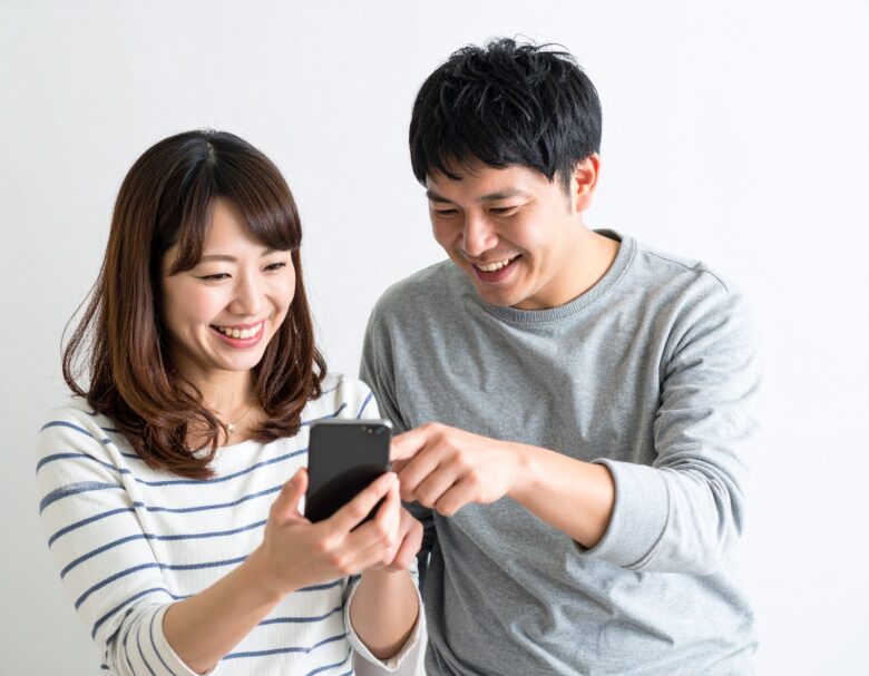 日本人夫婦が一緒に携帯電話を見ていて、笑顔になっている。女性が携帯電話を持ち、男性がそれを指さしている