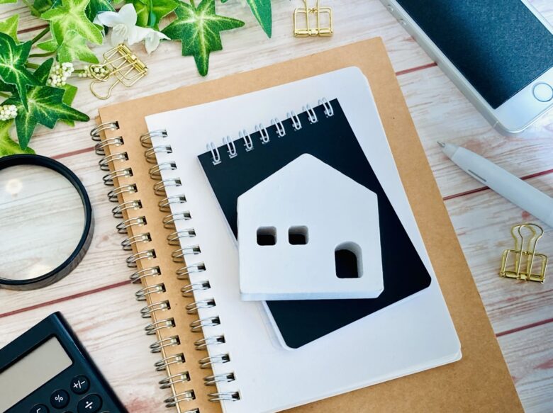 ノートが3冊重なっている上に白い住宅の模型が乗っている。周りには携帯電話や電卓、クリップなどが置いてある。